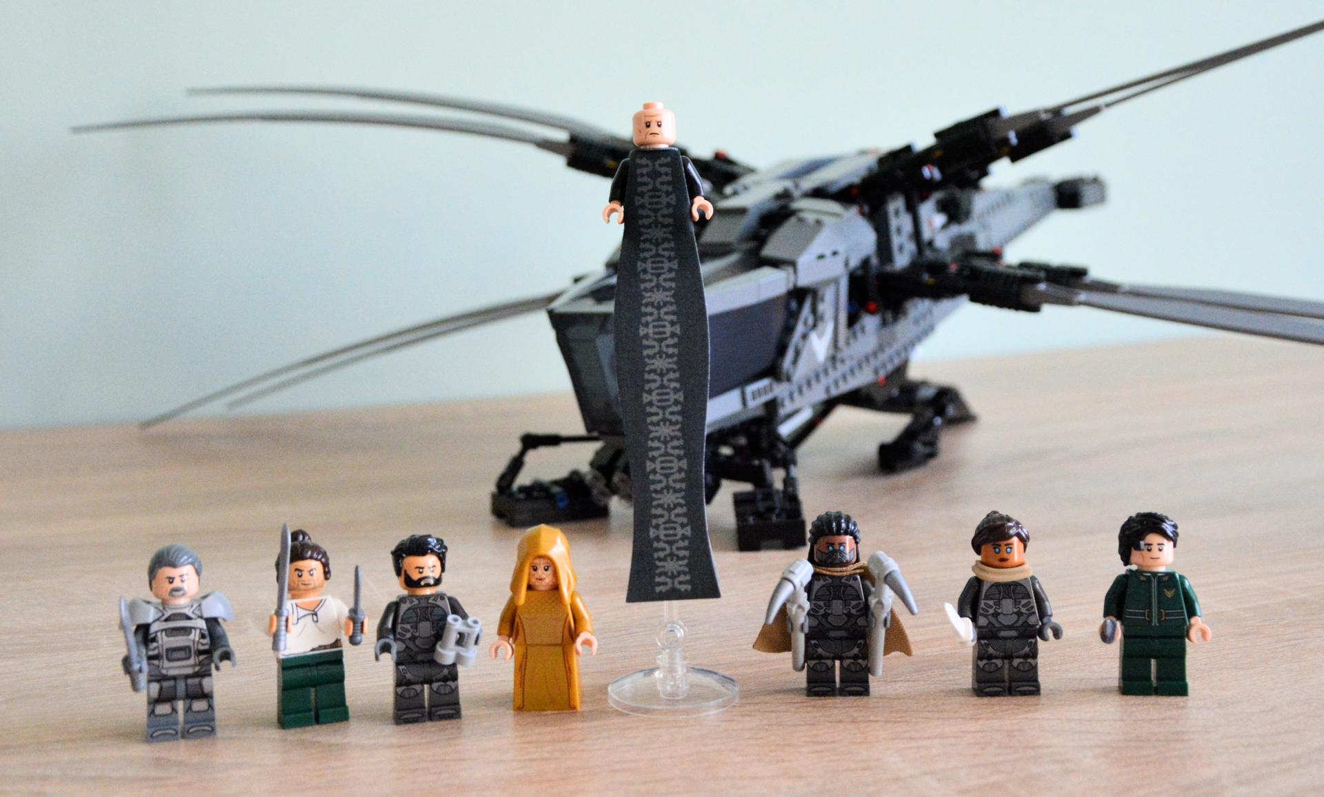 Na pierwszym planie stoi osiem figurek Lego przedstawiających
postacie z Diuny. W tle widać ornitopter z rozłożonymi skrzydłami.